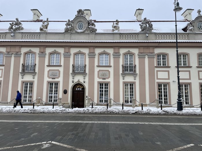 Historisk byggnad, barockdetaljer, skulpturer på taket, snö, person går, gatlykta, dagsljus, fönsterluckor, inristade dekorationer.