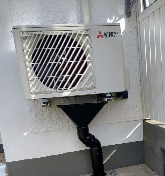 Vit luftkonditioneringsenhet av Mitsubishi Electric monterad på en vit vägg utomhus med svarta rördetaljer under.