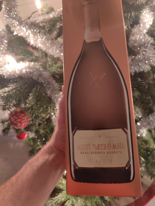 Förpackning för mousserande vin framför julgran. Etikett säger "Agustí Torelló Mata Gran Reserva Barrica Cava". Festlig stämning.