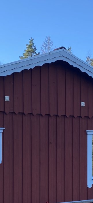 Rött trähus med vit takkant, ventilationsspalter, mot skog och klarblå himmel.