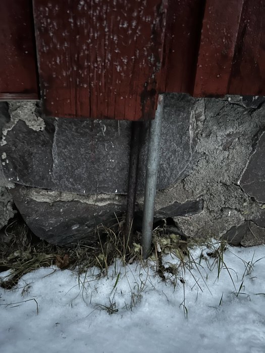 Rödfärgat trä, grå sten, metallstolpe, gult gräs, vit snö. Utomhus, detalj av byggnad, övergången vinter till vår.