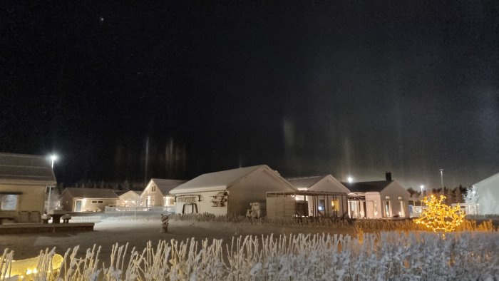 Ett snötäckt bostadsområde på natten med belysta hus och frostklädd vegetation, under en stjärnklar himmel.