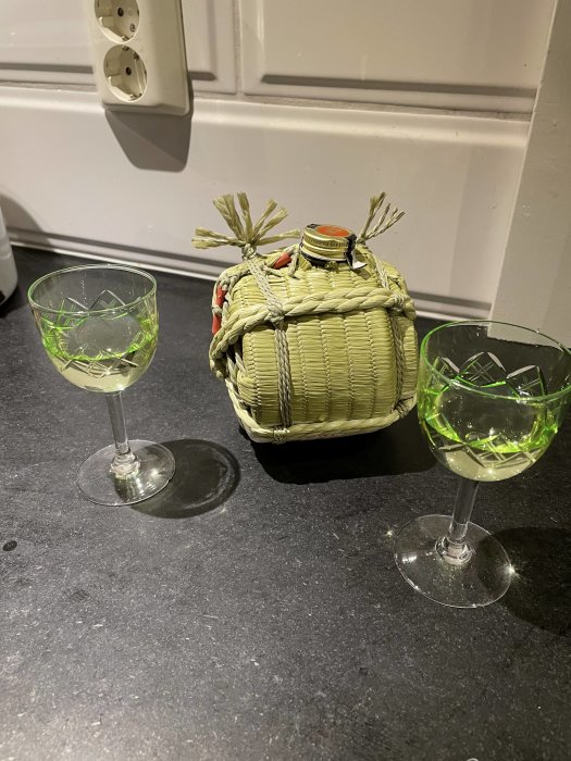 Flätad flaska omgiven av två glas med grön vätska på mörkt köksbord.