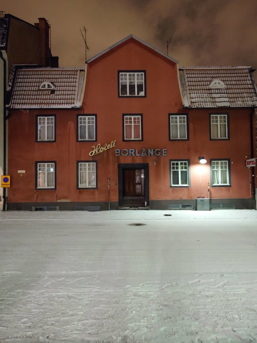 Tvåvåningshus i orange, snötäckt gata, skylt "Hotell BORLÄNGE", kvällsbelysning, tom gata, arkitektoniska detaljer, inget folk.