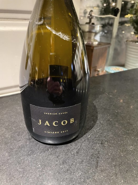 Vinflaska märkt "JACOB PREMIUM CUVÉE VINTAGE 2017" på köksbänk, julgran i bakgrunden.
