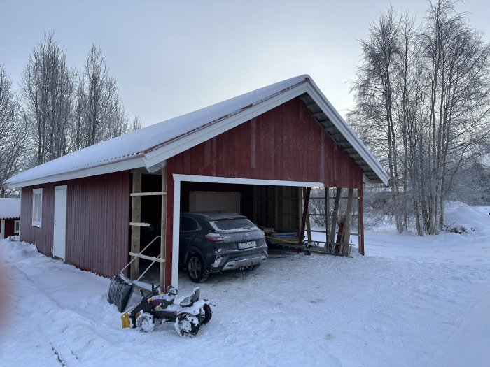 Röd byggnad med öppen garageport, delvis parkerad bil, snöslunga i snölandskap, träd, vinterdag.