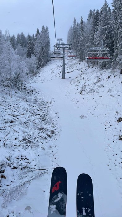 Skidåkarens perspektiv på en tom skidbacke med snötäckta träd och en lift, på en molnig dag.