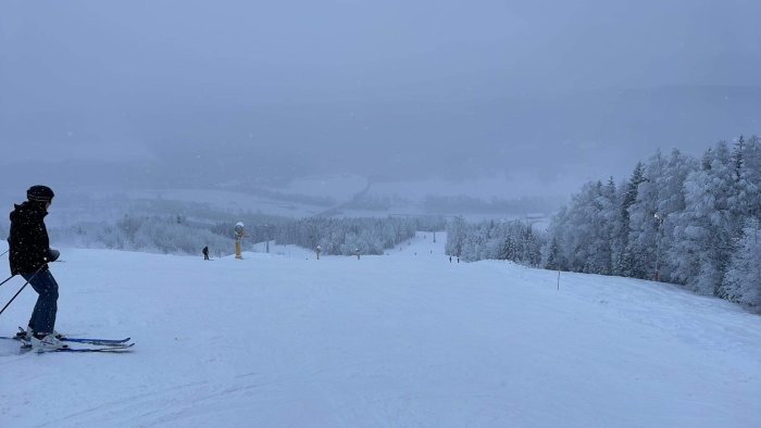 Skidbacke under snöfall med skidåkare, vinterträd och diffust landskap i bakgrunden. Dimmigt och kallt väder.