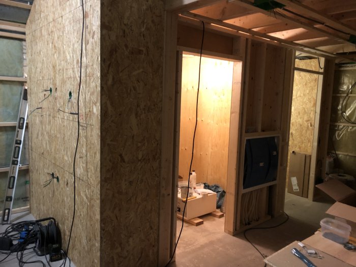 Ett pågående inomhusbygge med OSB-skivor, trästomme, isolering, stege och kablar. Oavslutat projekt, verktyg och byggmaterial syns.