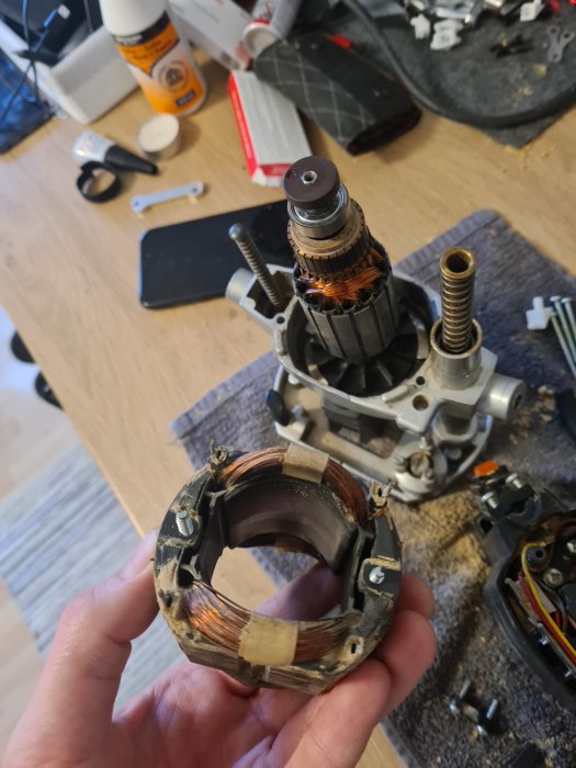 En hand håller en elektrisk motorstator framför en demonterad motor på ett bord med verktyg och delar.