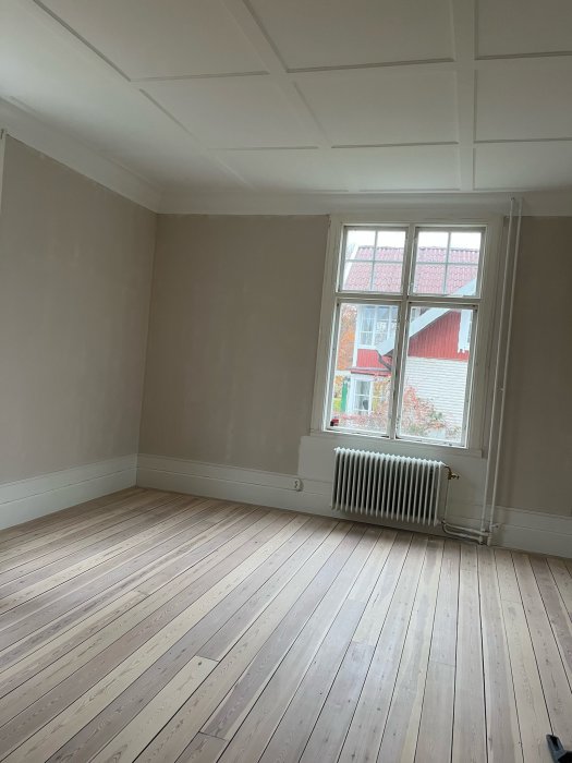 Tomt rum med trägolv, vita väggar, fönster och en radiator.