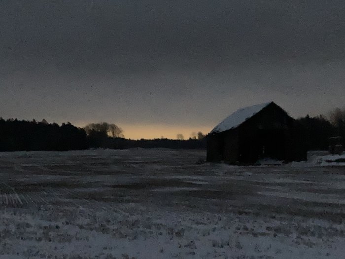 Mörkt vinterlandskap vid skymning, snötäckt fält, ensam lada, silhuetter av träd, dunkelt ljus i horisonten.