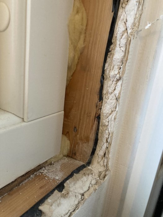 Skadad dörrkarm och väggisolering, exponerad trä och sprucken fyllning, under reparation eller skadekontroll.