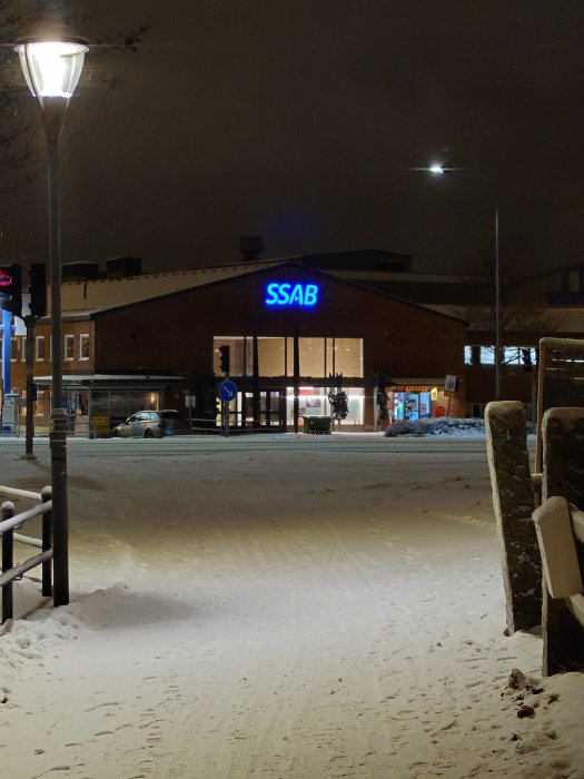 Nattbild av snötäckt gata med upplyst SSAB-skylt på byggnad och gatlyktor.