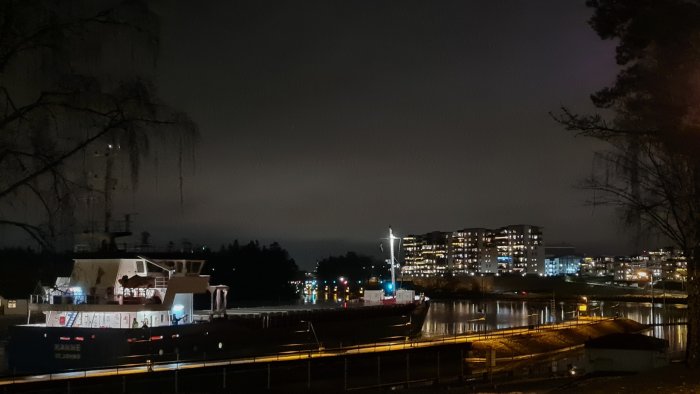 Natt, stadssilhuett, båt vid kaj, upplysta byggnader, molnig himmel, svagt ljus, reflektioner i vatten, mörk atmosfär.