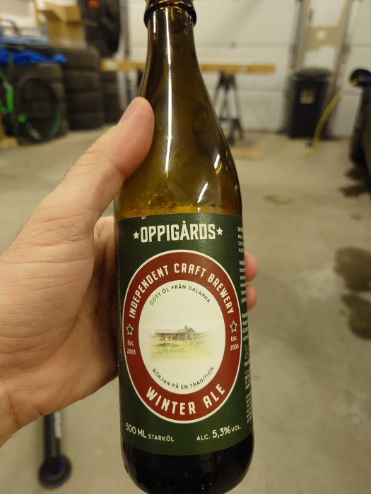 En hand håller en Oppigårds Winter Ale ölflaska; 5,3% alkohol, 500 ml, bryggeri från Dalarna, Sverige.