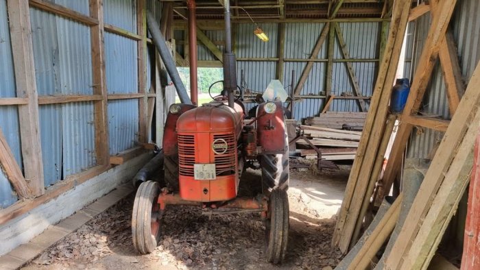 Gammal röd traktor inne i ett lantligt skjul med träbalkar och böjda plåtväggar.