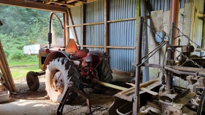 Gammal traktor i lada med jordbruksredskap, slitet, rustikt, lantligt, soligt utomhus genom öppning.