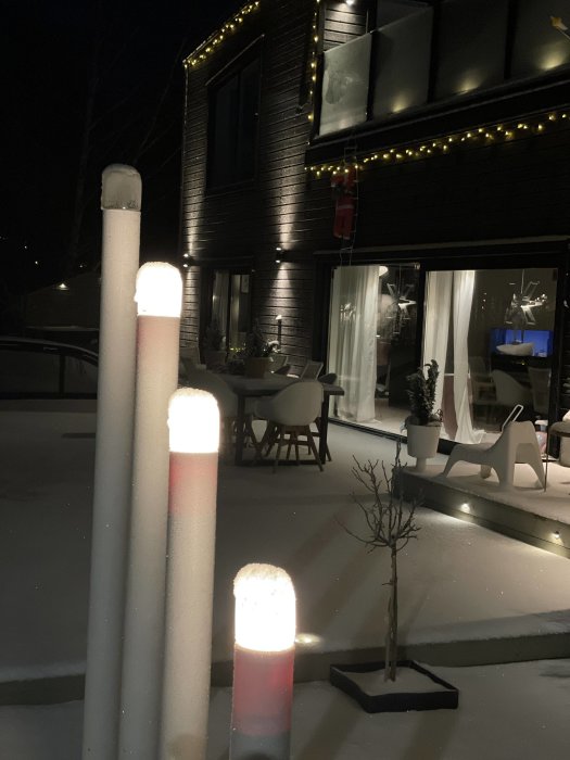 Snötäckt terrass med julbelysning, mörk byggnad, juldekorationer och upplysta stolpar i kvällsmörkret.