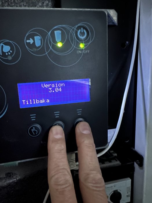 En hand trycker på knappar på en panel med LCD-skärm som visar texten "Version 3.04 Tillbaka".