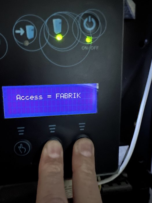 Maskinpanel med knappar, gröna lysdioder och LCD-visning med texten "Access = FABRIK". Två fingrar rör vid knappar.