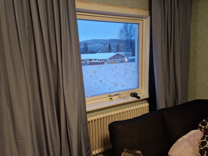 Vardagsrum med gardiner, fönster med utsikt mot snötäckt landskap, soffa, mönstrad rullgardin, radiator.