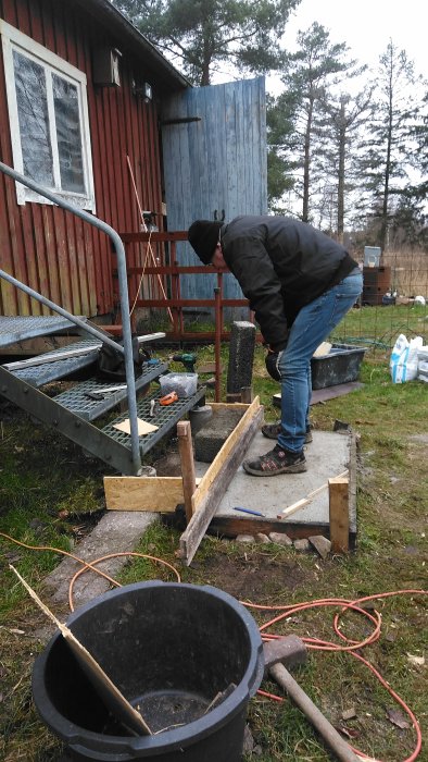 Person böjd över en utomhusgrill nära röd stuga, trädgårdsarbete och verktyg synliga, outvecklad utomhusmiljö.