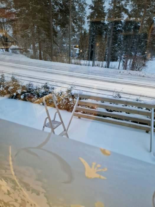 Vinterutsikt genom ett fönster, snötäckt landskap, träd, terassmöbler, handavtryck på fönstret.