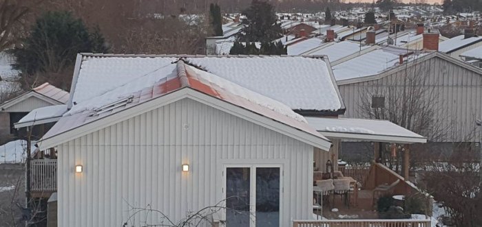 Ett vitt hus med snötäckt tak i ett bostadsområde under vinter. Terrass och träd i förgrunden.