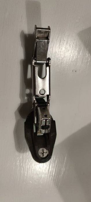 Metallisk skidhållare för sömnadsmaskin fäst på vit yta, närbild, skugga, sömnadstillbehör, vertikal orientering.