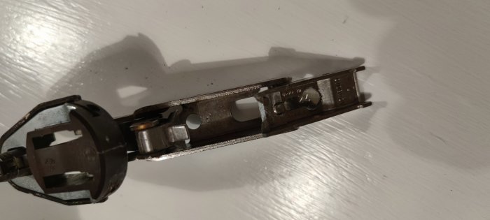 Metalliskt föremål, möjligen elektronik eller mekanisk del, på vit yta, detaljer synliga, komplex konstruktion, skruvar, metallplattor.