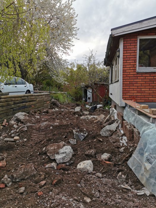 Husränna under renovering, stenar och jord, blommande träd, grå himmel, bil i bakgrunden, dagtid.