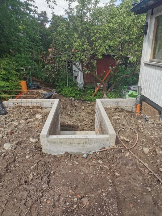 Byggarbetsplats med grävd grund och betongfundament, rörledningar synliga, grönska i bakgrunden, grumlig himmel.