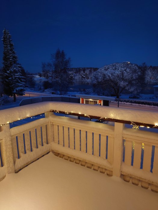 Veranda med snö, ljusslinga, vy över snötäckt landskap, träd, hus och sjö vid skymningen.