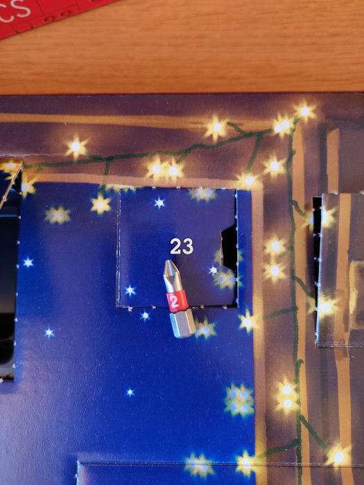 Adventskalender med öppnat lucka nummer 23, visar en leksakskula, stjärnmönster i bakgrunden.