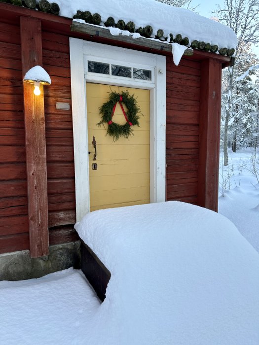 Röd stuga, gul dörr, julkrans, snötäckt, vinter, belysning, tyst, kallt.