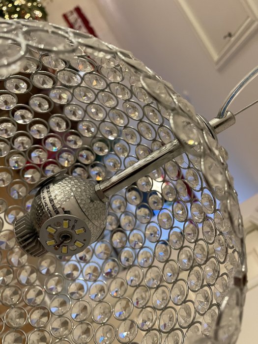Närbild av en kristallkrona med LED-lampa, genomskinliga prismor, metallram, oskarp bakgrund med juldekorationer.