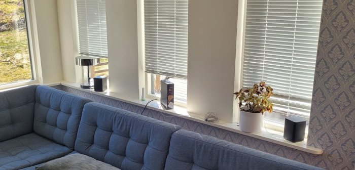 Ett vardagsrum med soffa, fönster med persienner, plantor och teknikprylar på fönsterbrädan i solljus.