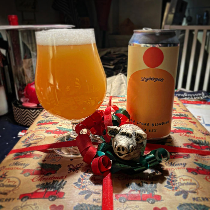 Ölglas fyllt med grumligt öl bredvid burk, grishuvud med julklappsband, julduksmönster.