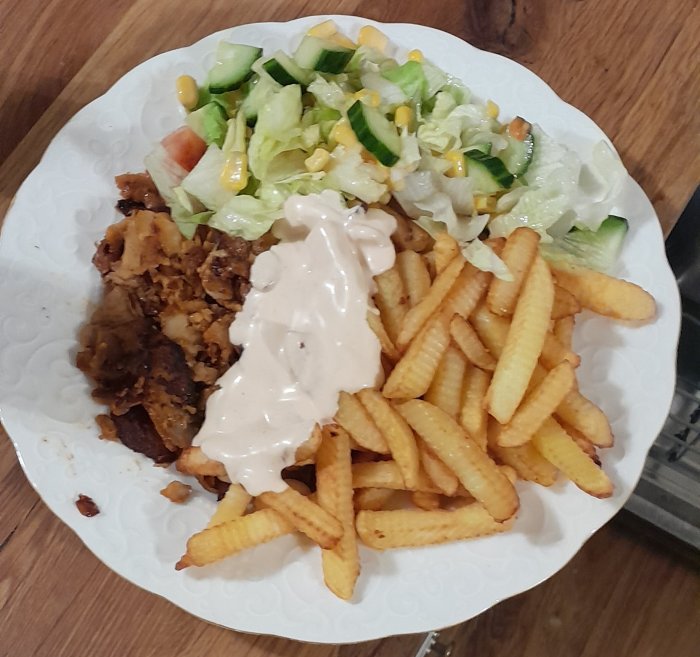 En tallrik med sallad, pommes frites och friterad kyckling täckt med sås, serverad på träunderlag.