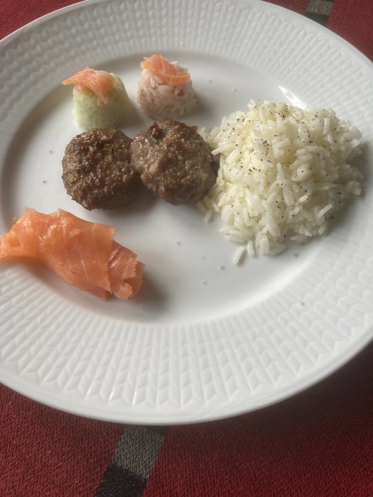 Vitt ris med svartpeppar, köttbullar, sushirullar med lax på ett vitt fat.