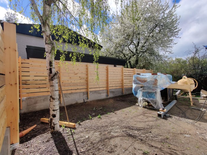Trädgård under konstruktion med staket, träd, cementblandare och byggmaterial, delvis soligt väder.