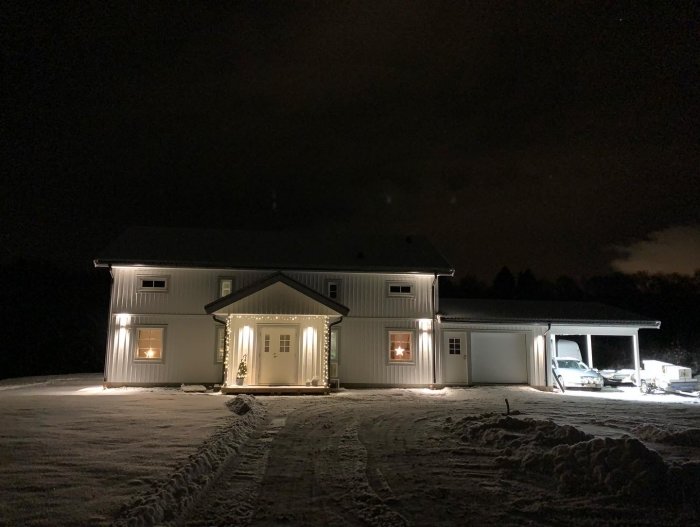 Ett vitmålat hus omgivet av snö, upplyst på natten, vinteratmosfär, parkerade bilar i carport.