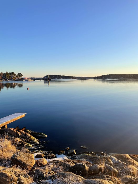 Stilla vatten, brygga, klippor, lantligt landskap, klar himmel, tidig morgon eller skymning, reflektioner, fridfullt.