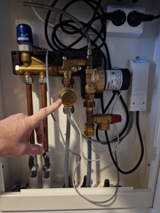 Person pekar på VVS-installation med rör, ventiler, och kablar i ett skåp.