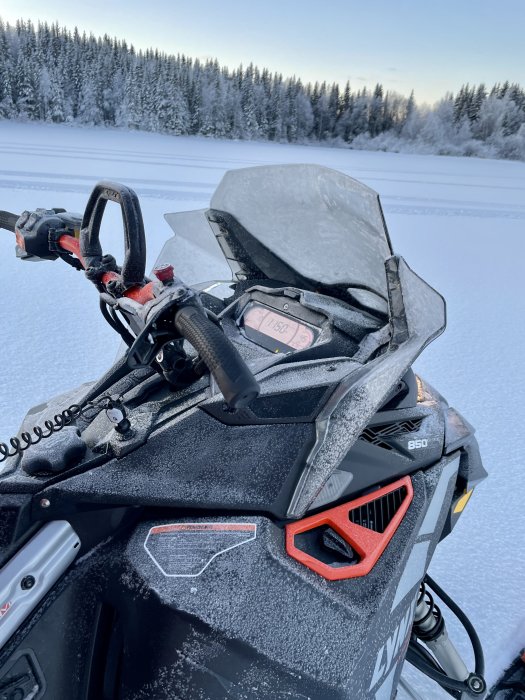 En snöskoter täckt av frost mot en vintrig landskapsbakgrund med snöklädda träd och klar himmel.