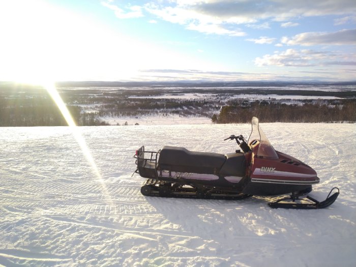 Snöskoter på vinterlandskap, snötäckt mark, träd i fjärran, solnedgång med solstrålar, klar himmel.