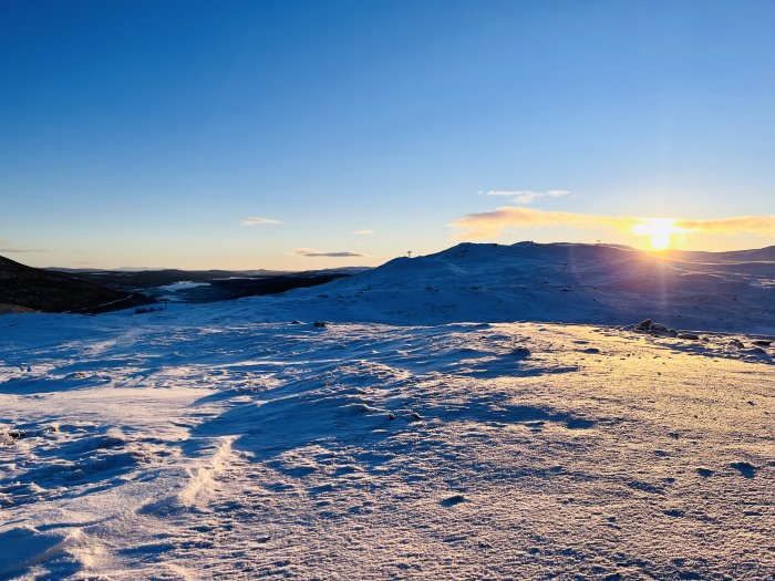 Solnedgång, snötäckt terräng, vinterlandskap, klar himmel, skidliftar i fjärran, fredlig, spegling av solljus på snön.