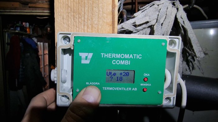 En hand håller en grön värmeregulator märkt "THERMOMATIC COMBI" monterad på en träyta i ett rörigt utrymme.