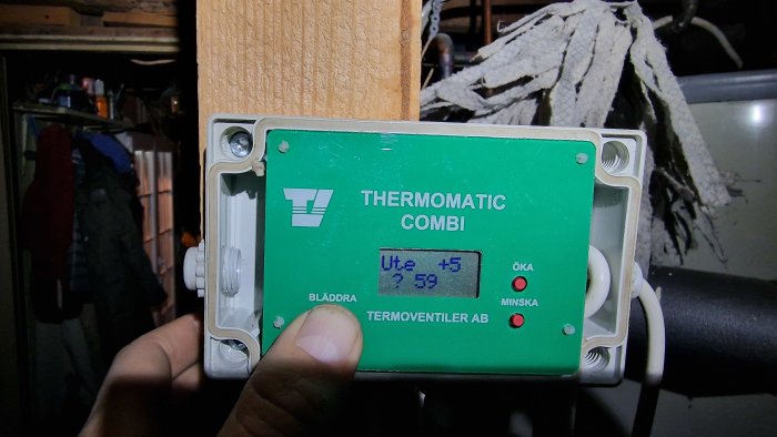 En person håller en elektronisk termostat vid en träbalk. Apparaten är märkt "THERMOMATIC COMBI" och har LCD-display.